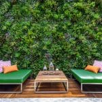 A modern outdoor seating area features two green cushioned benches with colorful pillows. A wooden coffee table holds decorative lanterns. Behind, a lush vertical garden wall with various green plants creates a vibrant backdrop.