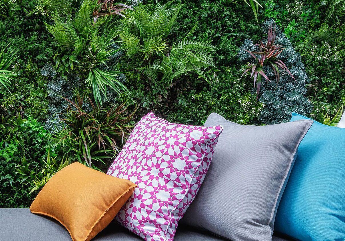 A collection of colorful cushions in shades of pink, gray, blue, and orange are placed on a sofa. Behind them is a lush vertical garden with a variety of green plants and ferns, adding a natural backdrop to the scene.