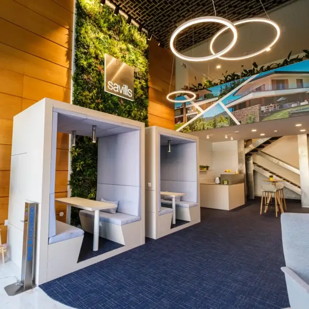 Modern office space with blue pods for private meetings, green wall decor, circular light fixtures, and a branded Savills sign. Upstairs area with a desk and bar stools visible. Warm wooden walls complement the sleek design.