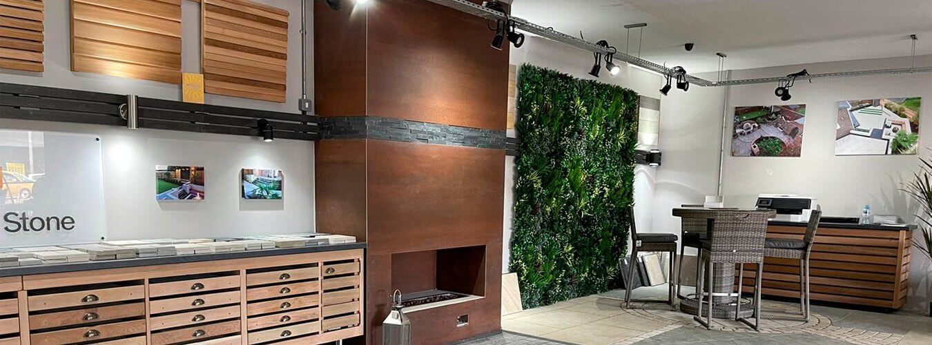 A modern interior showroom featuring wooden drawers with samples, a fireplace with brown panels, a wall with green plants, and a corner with a small table and chairs. Ceiling track lights illuminate the space, and photos decorate the walls.