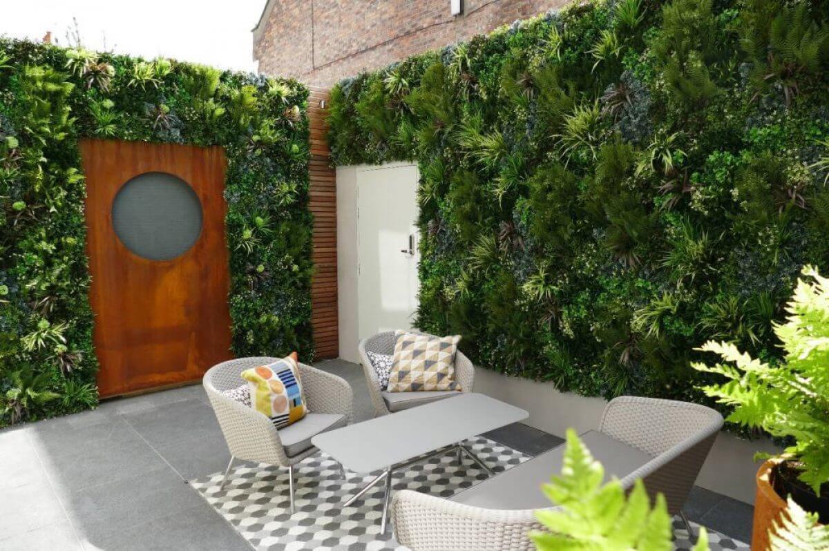 Modern outdoor patio with gray wicker chairs and a low table on a checkered rug. Vertical green plant walls surround the space. A wooden door with a circular window is visible, and a variety of cushions add color to the seating area.