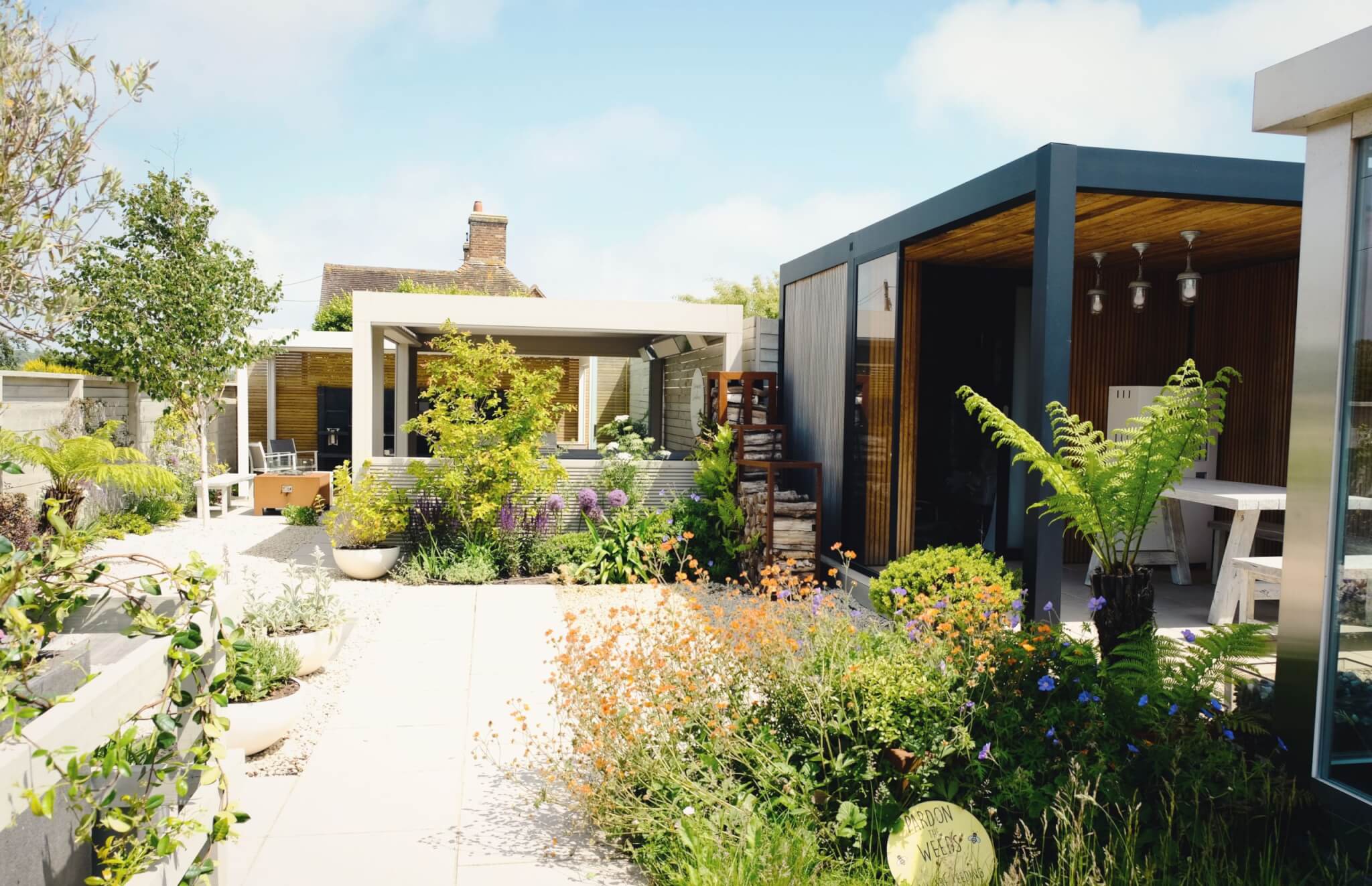 A modern garden with two outdoor structures surrounded by lush greenery and blooming flowers. The patio features a mix of plants and flowers, including tall ferns and brightly colored blooms, under a clear blue sky.