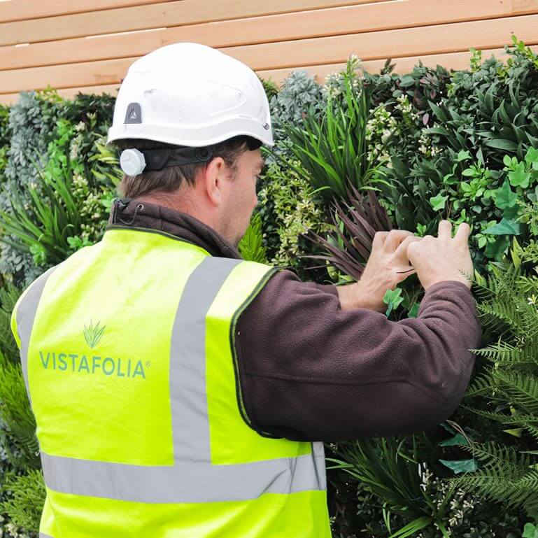 A person wearing a yellow vest with VistaFolia on the back and a white hard hat is adjusting an artificial green wall adorned with various plants. Wooden panels are situated above the vibrant display.