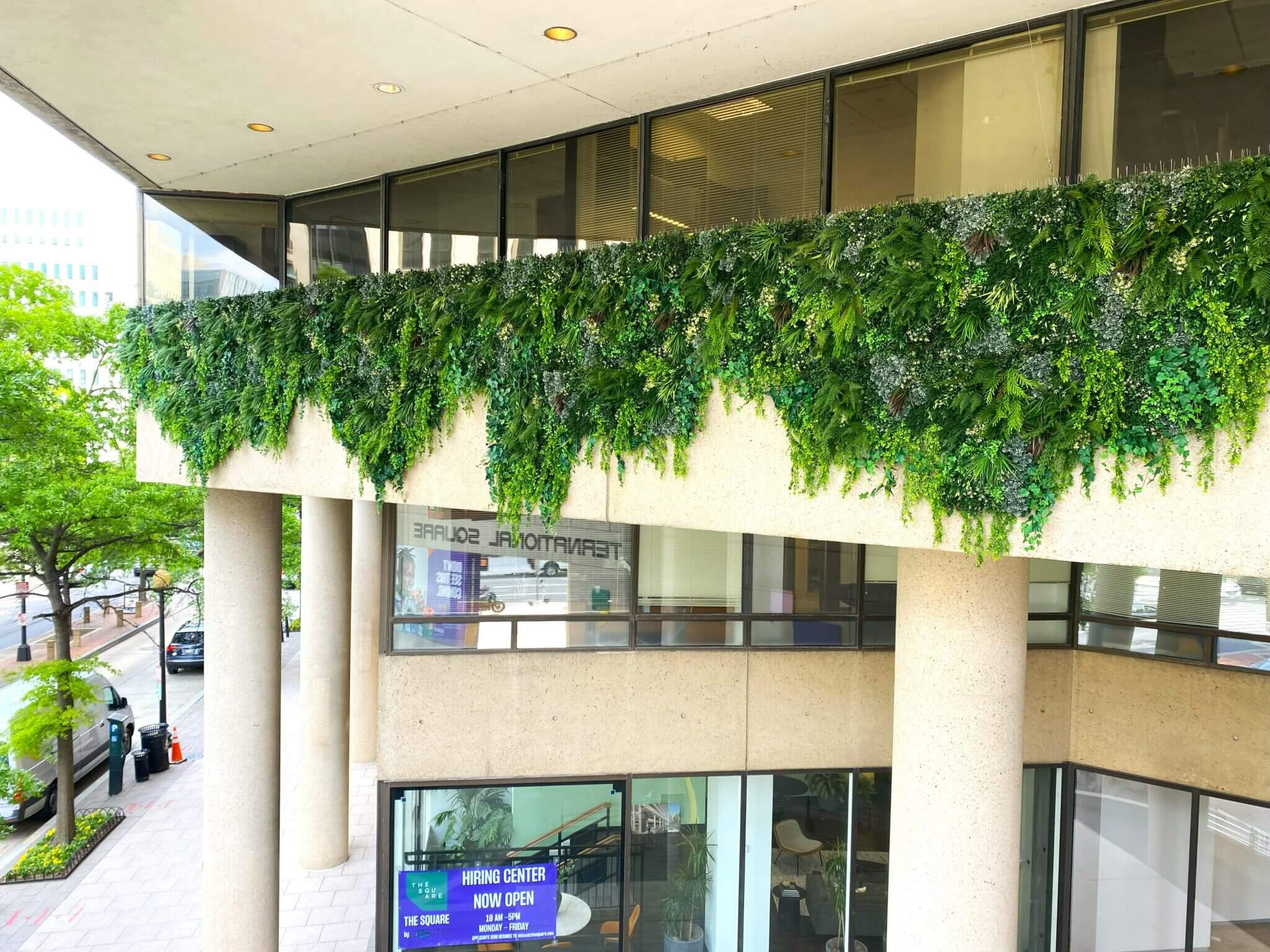 A commercial office block in Washington DC features a lush green vertical garden on its upper level. Large windows and concrete pillars enhance its modern design. The street view shows trees and cars passing by, while a sign indicates the leasing center is open.