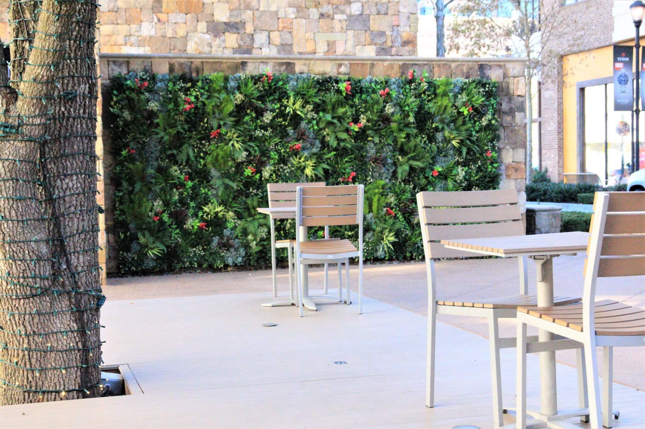 Outdoor patio with wooden tables and chairs, set against a stunning outdoor faux green wall in Alpharetta, Georgia, adorned with vibrant red flowers. A tree wrapped in string lights stands elegantly in the foreground. Stone wall and walkway add to the charming backdrop.