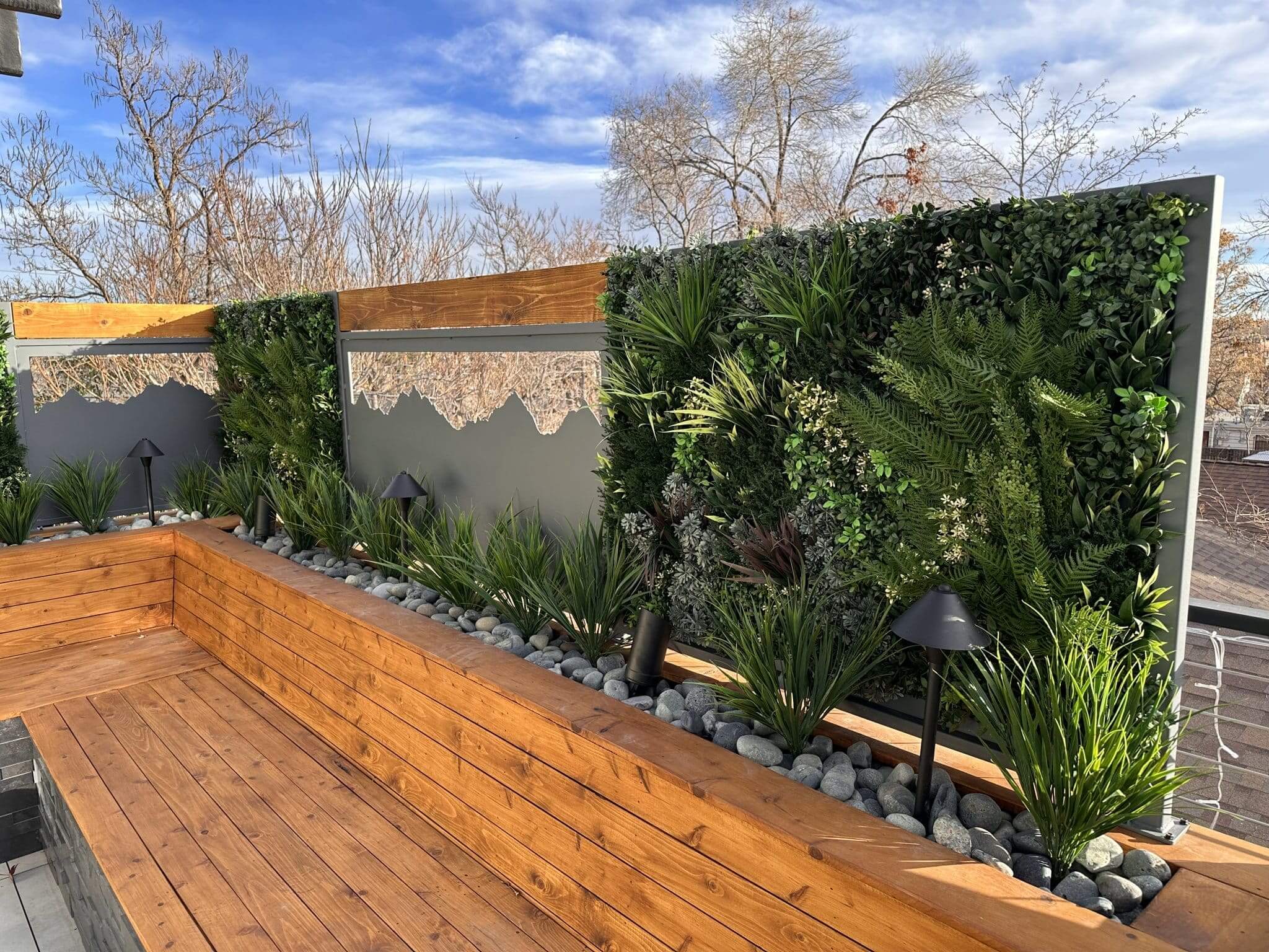 A rooftop garden in Denver showcases wooden benches, decorative panels, and a lush green wall. The area is adorned with round stones and modern black lamps against a backdrop of bare Colorado trees and a blue sky.