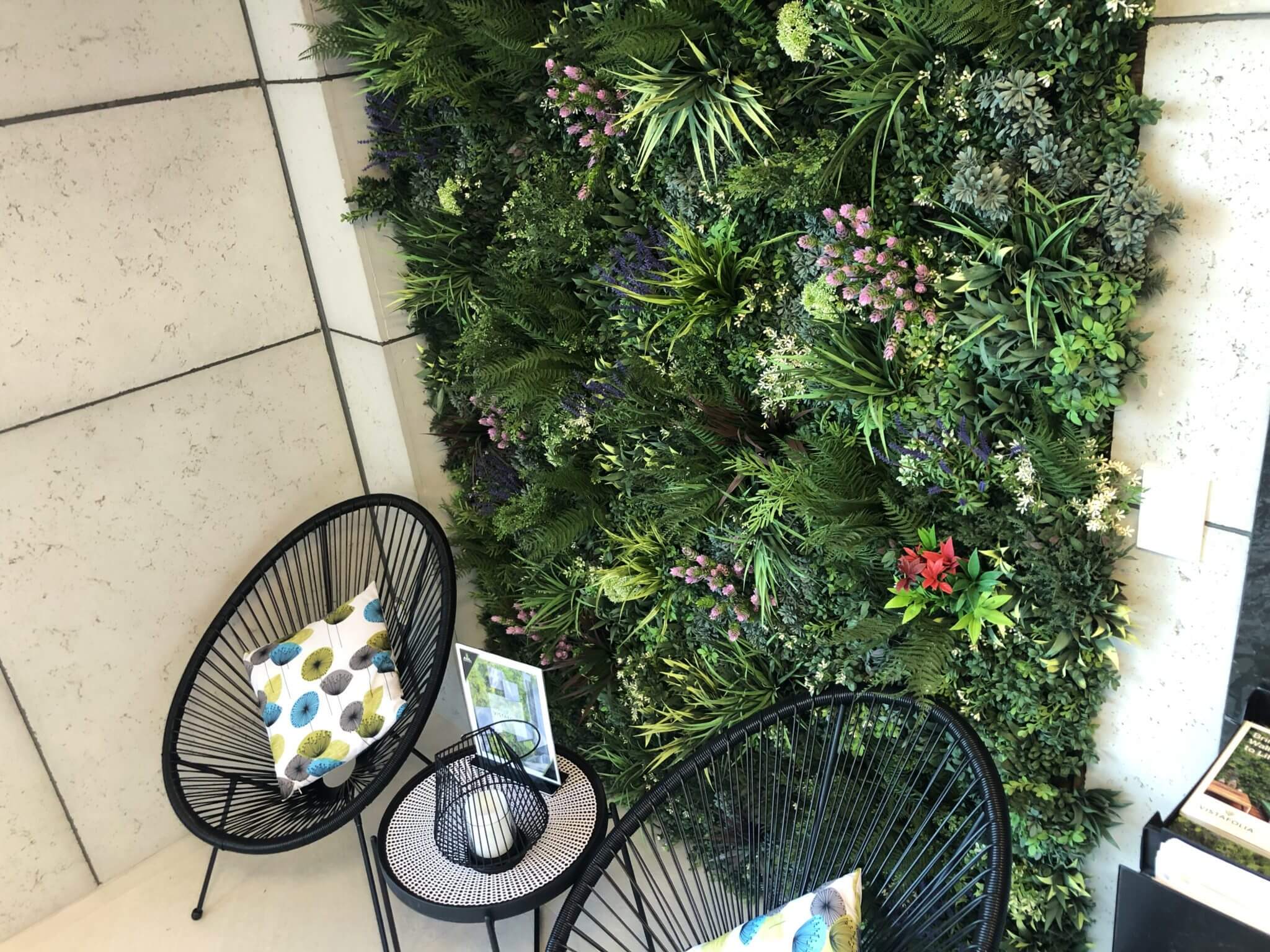 Indoor seating area with a lush vertical garden on the wall. Two black wire chairs with patterned cushions are arranged around a small round table. The garden features various green plants and colorful flowers.