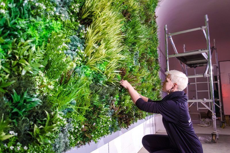 A person with short hair tends to a lush vertical garden indoors, adjusting plants on a wall covered in greenery. A scaffold is nearby, suggesting ongoing work.