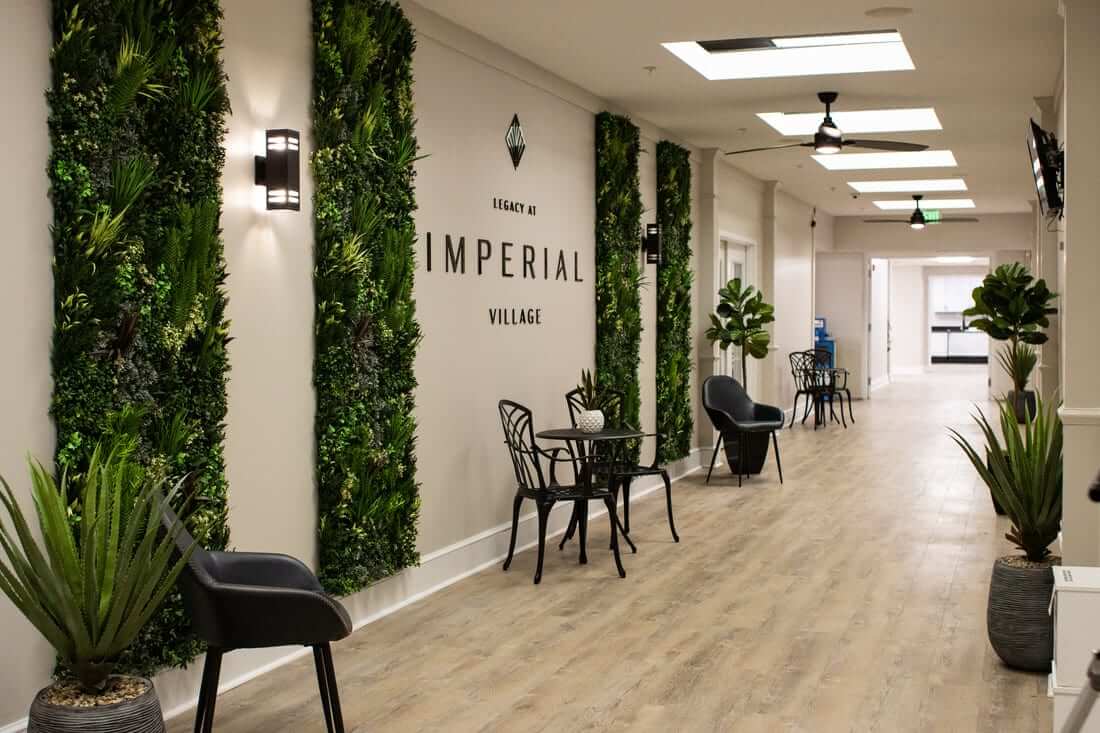 A modern hallway with wooden flooring boasts a vibrant Care Home Green Wall in Richmond, Virginia, alongside black chairs and small tables. The wall reads Legacy at Imperial Village, as natural light filters in from skylights above.