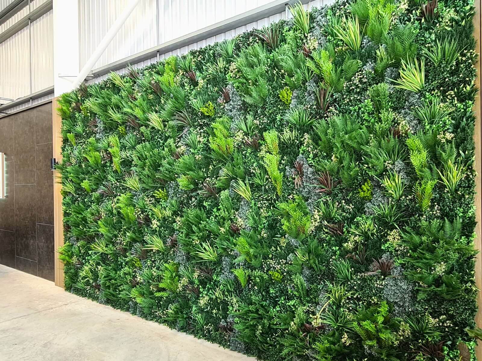 A large green vertical garden wall filled with various lush plants and foliage in an indoor setting, featuring a variety of textures and shades of green. The wall is adjacent to a concrete floor and a dark tiled area.