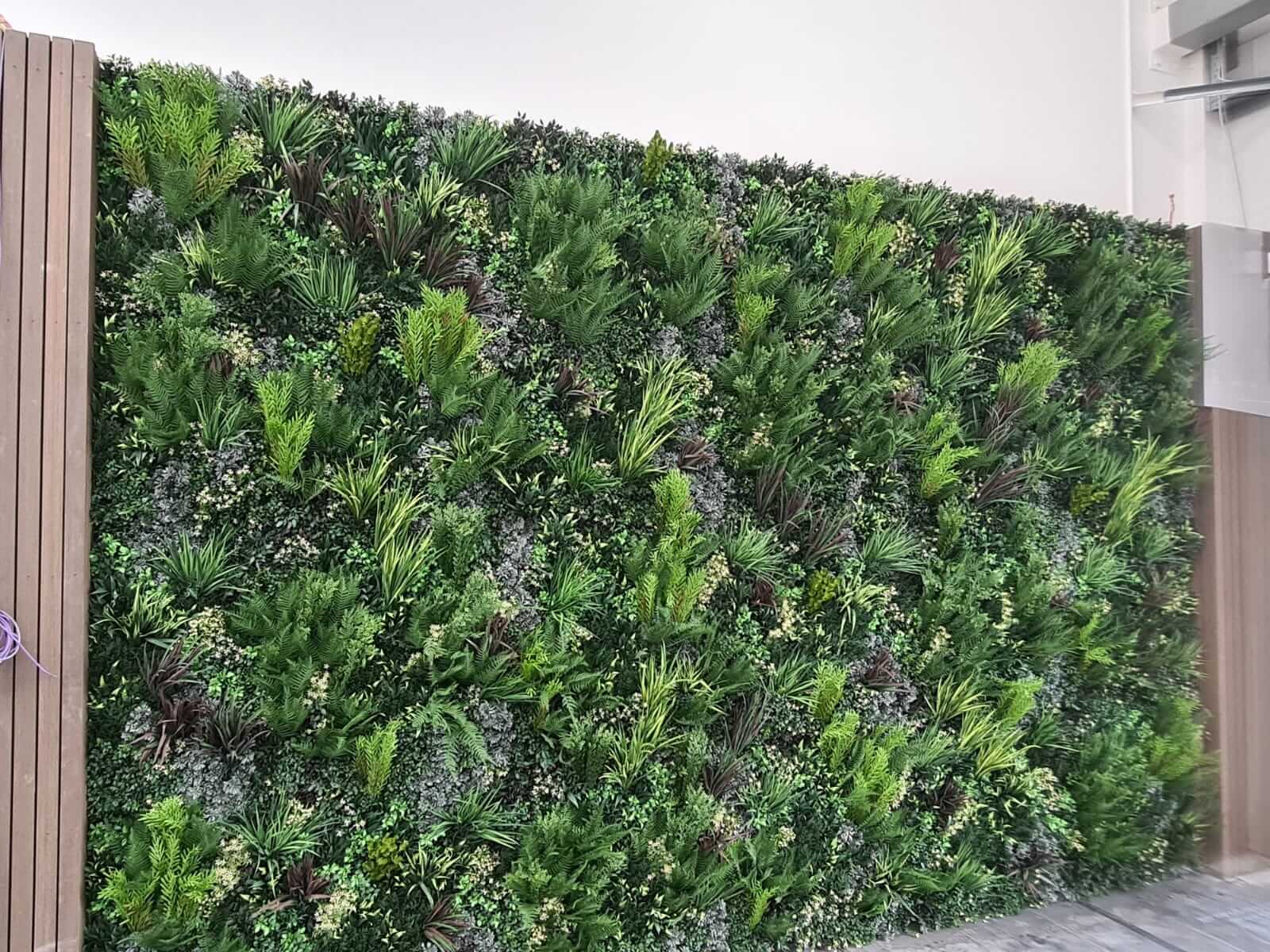 A lush vertical garden wall indoors, featuring a dense variety of green plants and foliage, creating a natural and vibrant atmosphere. The wall is framed by wood panels on the sides.