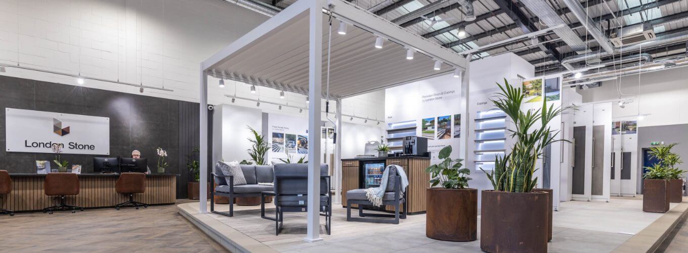 A modern interior showroom featuring a pergola with seating underneath. The space includes potted plants, wooden flooring, and display panels on the walls. Theres a reception area with a sign that reads London Stone in the background.