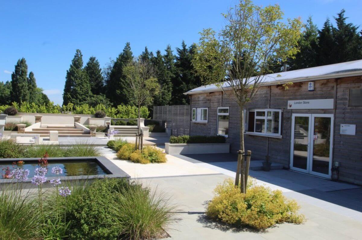 A serene outdoor area featuring a modern wooden building, surrounded by lush greenery and small trees. A rectangular water feature with floating flowers is in the foreground, under a clear blue sky.