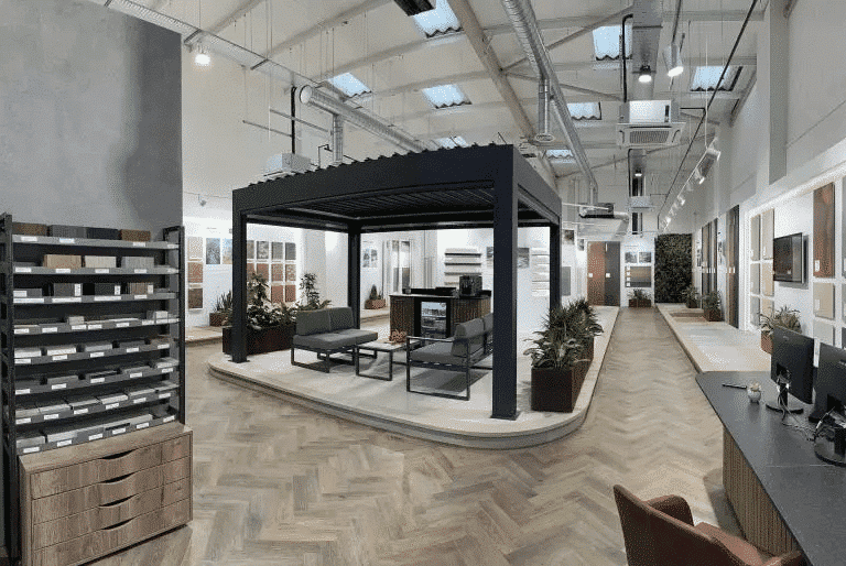 A modern showroom featuring a pergola display with seating and plants. The space has a variety of wall samples, large windows, and a sleek counter with computers. The floor is covered in a herringbone pattern.