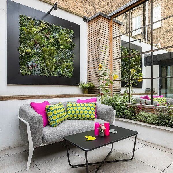 A stylish and modern outdoor patio features a gray sofa with vibrant pink and yellow pillows. A black coffee table holds pink candles. A green wall garden design adds life to the space, while sunflowers are reflected in a large mirror.