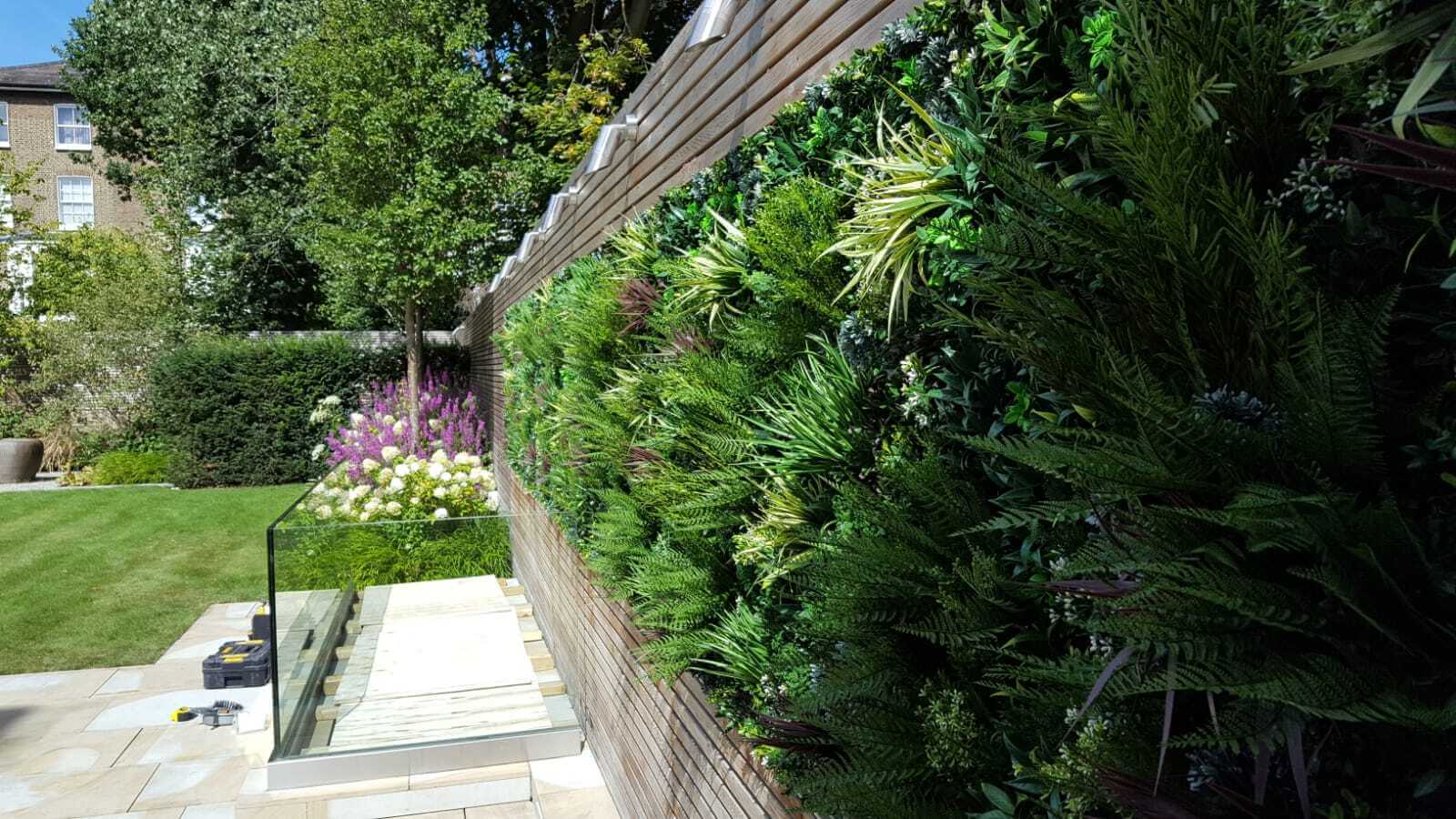 A modern garden with a lush vertical green wall on the right, featuring various plants. In the background, theres a manicured lawn, colorful flowerbeds, a glass railing, and steps leading to a lower area. Trees and a building visible in the distance.