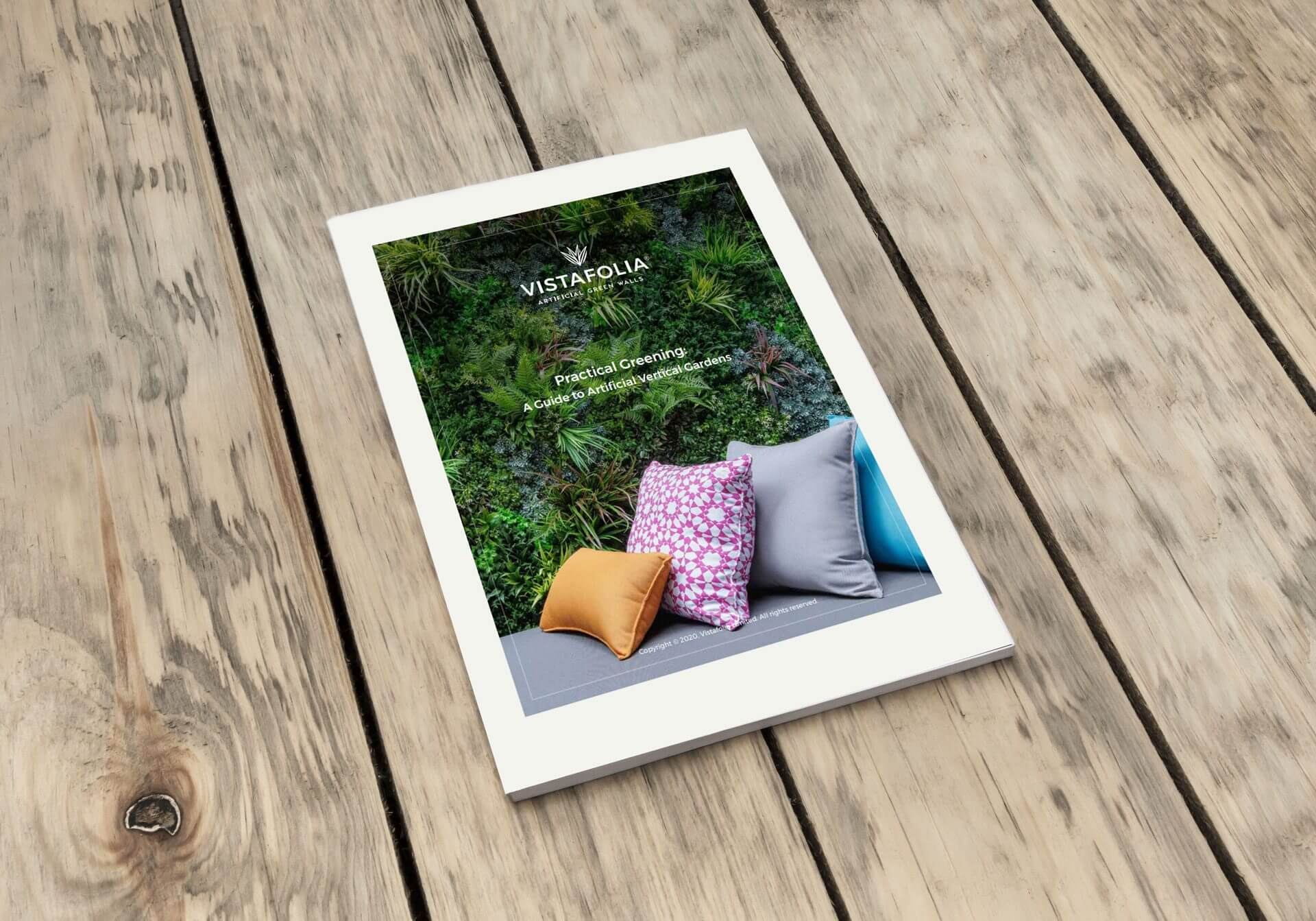 A brochure with a green plant wall on the cover, featuring colorful cushions in orange, pink, and gray. It is titled Vistafolia Practical Greening. The brochure is placed on a wooden surface.