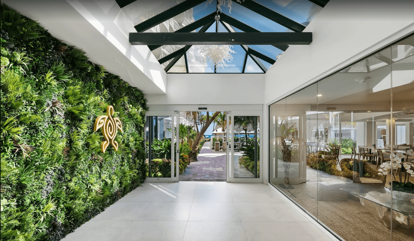 A modern indoor space with a glass ceiling in a Florida hotel features a vibrant green living wall and a gold turtle emblem. To the right, there's a glass-walled room with decorative plants. Glass doors lead outside to a brick pathway lined with lush greenery in Pompano Beach.