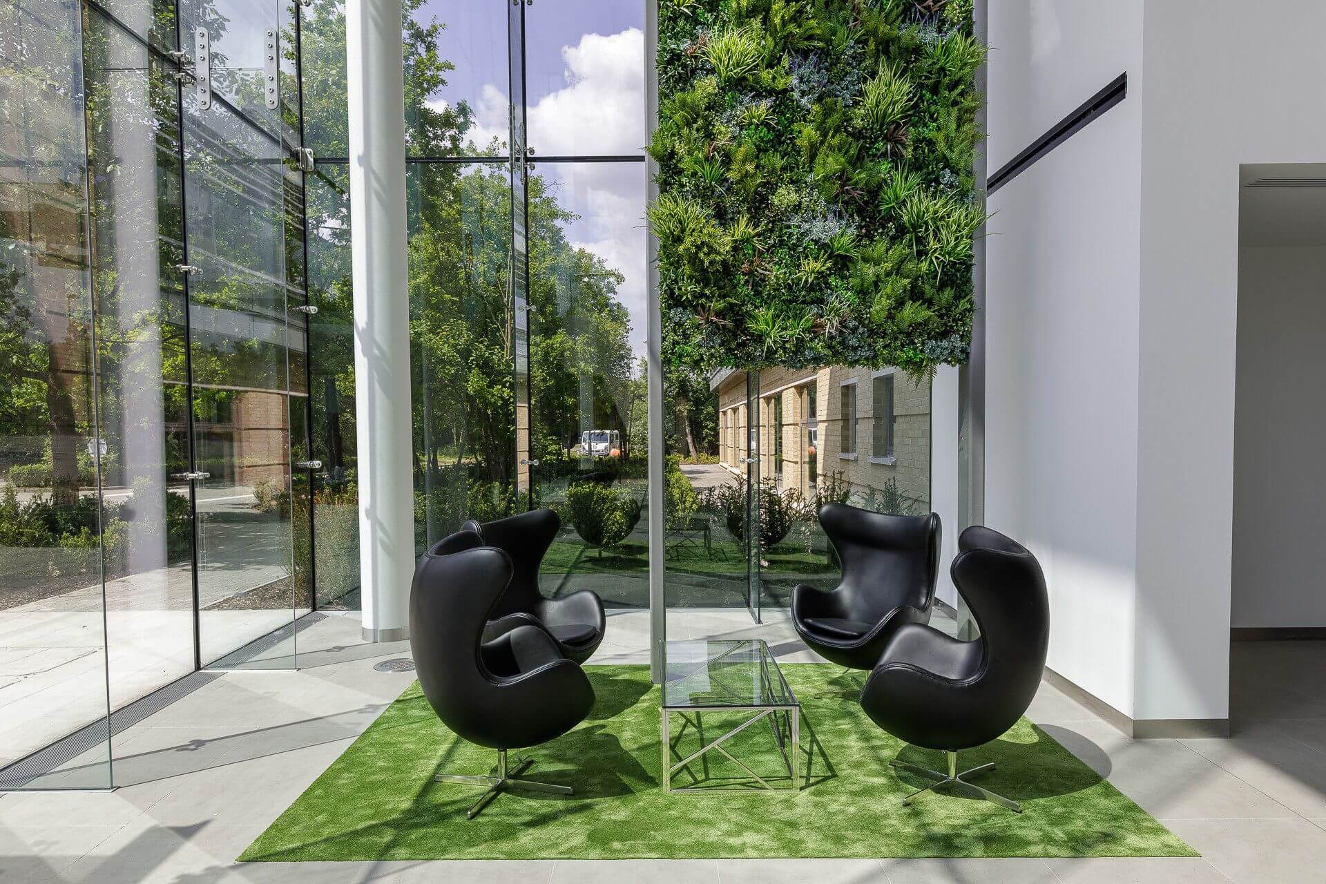 A modern sitting area with four black chairs around a glass table on a green rug. A vertical garden adorns the wall. Large windows offer a view of trees and a building outside.