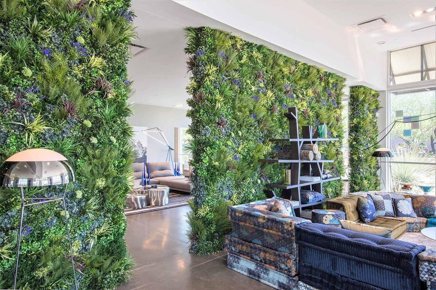 A modern living space featuring lush green walls with Vistafolia Green Wall Panels. The room showcases contemporary furniture, including blue sofas, a floor lamp, and a metal bookshelf. Large windows flood the area with natural light, enhancing the fresh, vibrant ambiance.