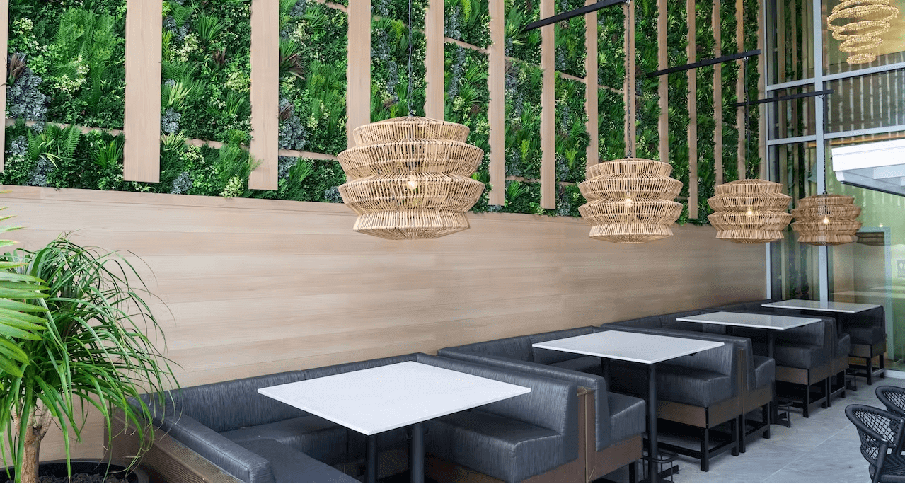 A modern restaurant interior featuring a vertical green wall, natural wood paneling, and several booth-style tables. The seating boasts dark cushions beneath woven rattan pendant lights, while an artificial potted plant adds a touch of greenery.