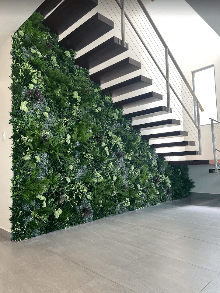 A modern residential interior in San Juan, Puerto Rico, showcases a staircase with a vibrant faux living wall underneath, filled with various plants. The space is bright and minimalistic with light-colored flooring and sleek metal railings.