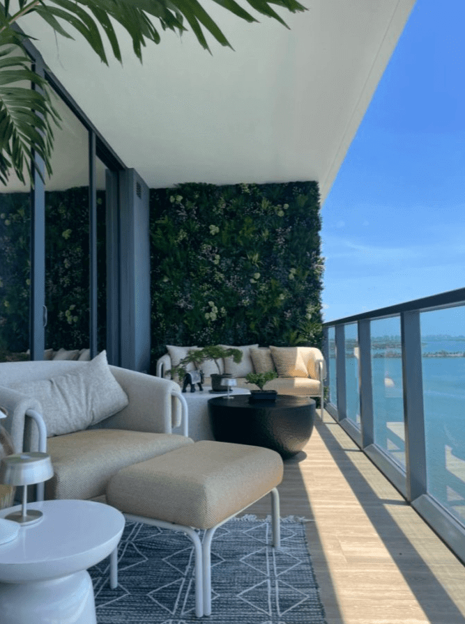 A modern balcony in Miami, Florida, features beige seating, a small table, and an artificial green living wall. The blue sky and expansive water view create a serene atmosphere. A potted plant adds greenery, enhancing the tranquil setting.