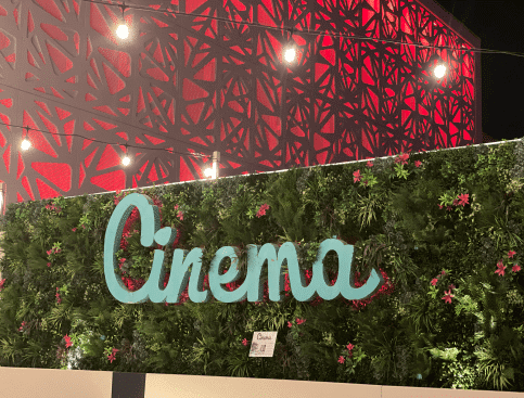 A decorative wall with lush green foliage, known for its hospitality faux green wall backdrop in Scottsdale, features the word Cinema in large, cursive blue letters. The Arizona backdrop includes intricate red patterns and string lights, creating a festive atmosphere.