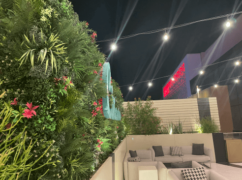 A rooftop patio at night in Arizona features a lush, Hospitality Faux Green Wall Backdrop in Scottsdale adorned with pink flowers. String lights hang overhead, illuminating outdoor seating with gray and white cushions. In the background, buildings are beautifully lit in red.