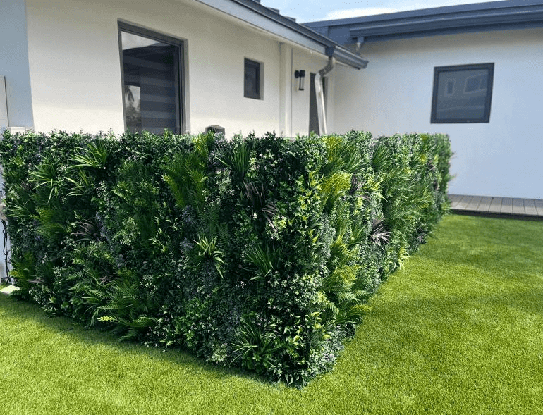 A neatly trimmed, lush green hedge surrounds a corner of a modern white house with a flat roof in Fort Lauderdale. Echoing the region's elegant garden design, the hedge sits on a vibrant lawn. The house features a sliding glass door and windows that blend indoor living with the outdoors seamlessly.