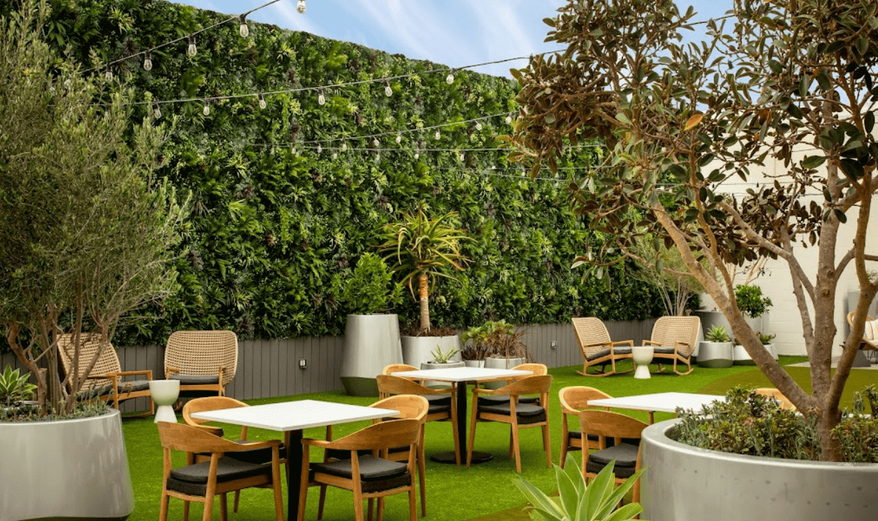 A modern outdoor patio at Hotel Green Wall in San Diego features tables and chairs on artificial grass, surrounded by planters and a lush green wall. String lights are hung above, creating a cozy California atmosphere.