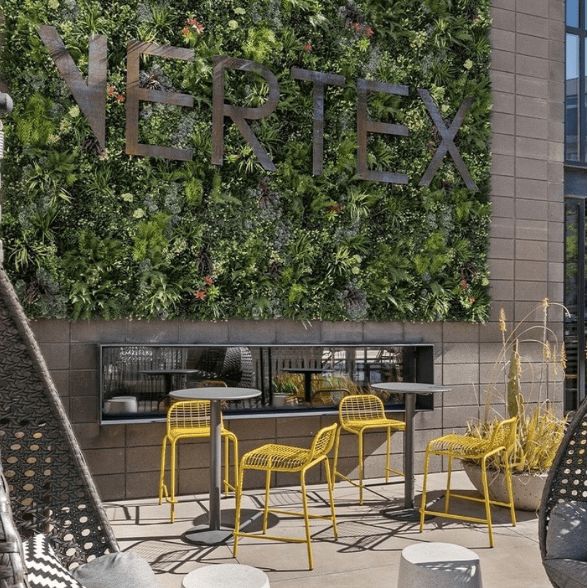 Outdoor patio with two yellow chairs and a table set against a wall with a vertical Multi-Family Green Wall in Phoenix. The word VERTE is visible on the wall. Large wicker furniture and decorative plants are present, creating a relaxed Arizona atmosphere.