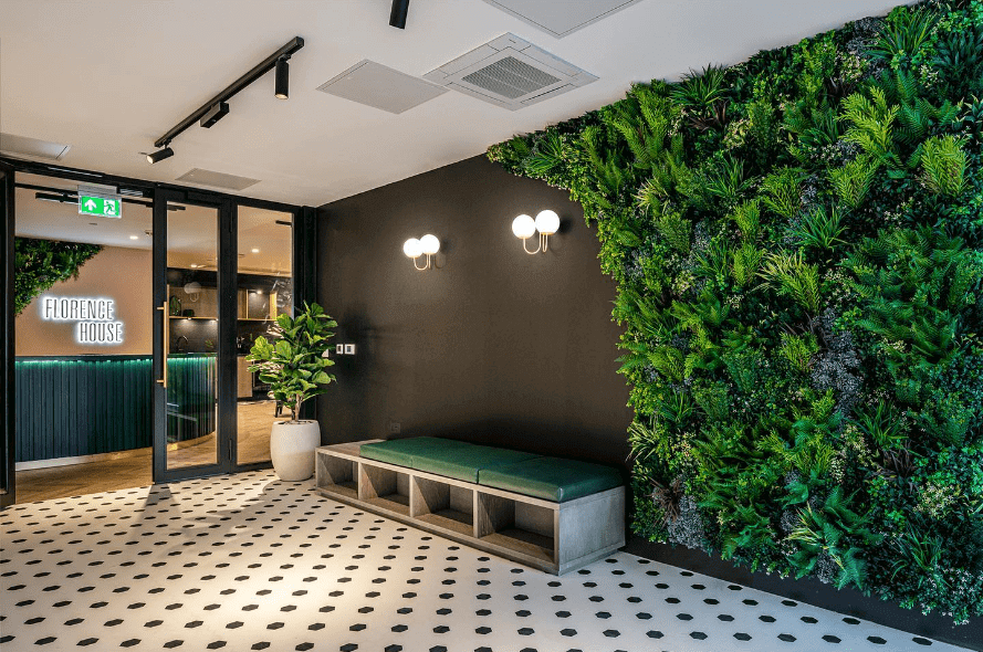 The modern lobby features an artificial plant-covered green wall, black ceiling lights, a green cushioned bench, and a black and white patterned floor. A sign reads Florence House near the reception area adorned with indoor plants.