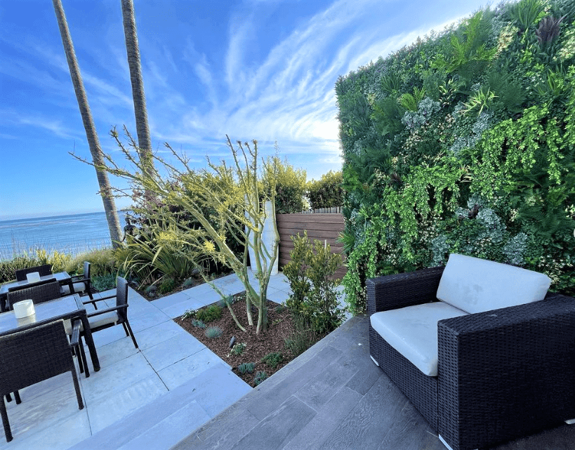 Outdoor seating area with wicker furniture and a white cushion, set near the ocean on a seafront property in California. A vertical garden wall and a small tree enhance the greenery. A few chairs and tables are on a tiled patio under a clear blue sky in Pismo Beach.