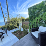 A serene outdoor patio at a seafront property in Pismo Beach, California, features wicker furniture overlooking the ocean. A white cushioned chair rests beside a lush green wall. A table with chairs is positioned closer to the sea, surrounded by tall palm trees and vibrant greenery.