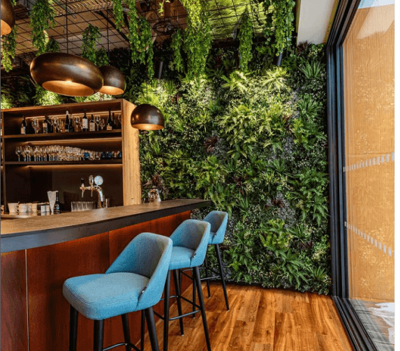 A contemporary bar area showcases a wooden counter paired with teal bar stools. Brass pendant lights hang overhead, casting a warm glow on the space. The back wall is beautifully decorated with Texture Foliage, forming an eye-catching vertical garden. Large windows on the right flood the area with natural light.