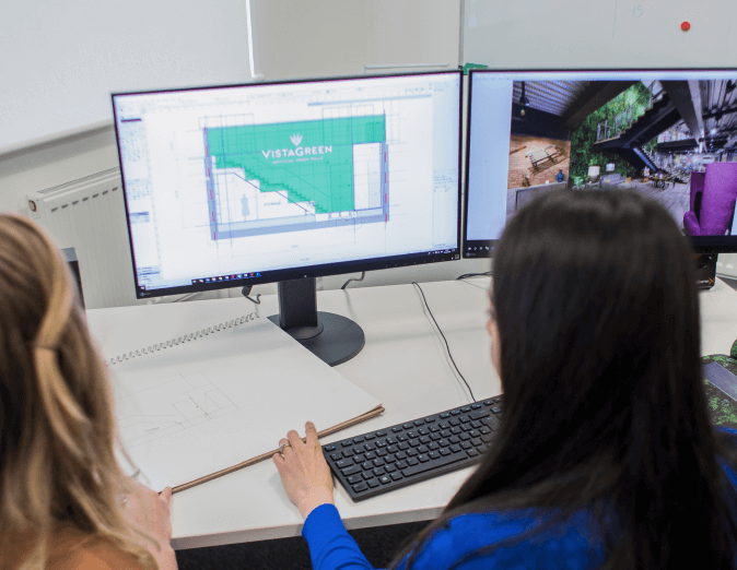 Two people work at a desk with two monitors showing architectural designs. One screen displays a layout with the text VISTAGREEN, while the other shows an interior space concept. A sketchbook with drawings is on the desk.