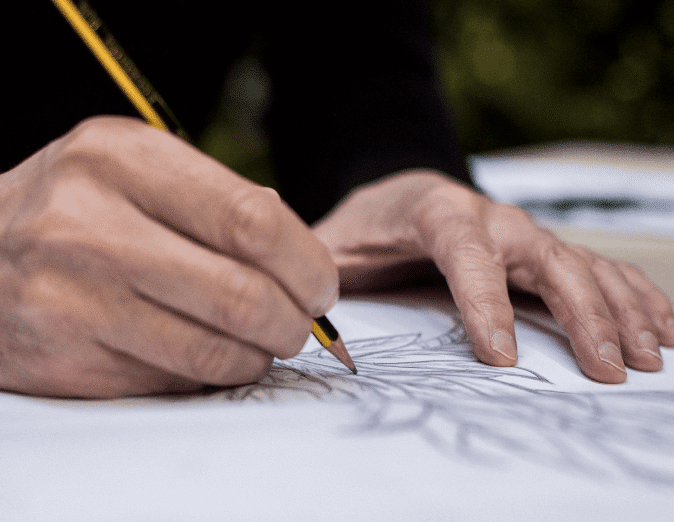 Close-up of hands sketching with a pencil on paper. The drawing appears to be floral with intricate details. The background is blurred, emphasizing the focused activity.