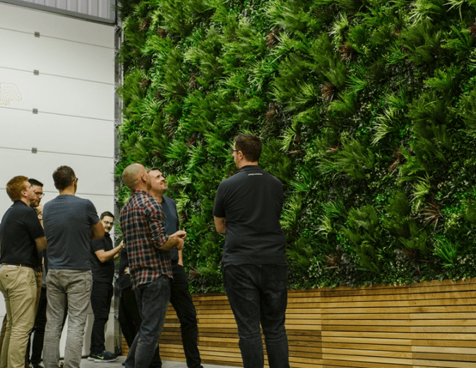 A group of people stand near a large, lush green wall made of various plants. They are engaged in conversation, some with their arms crossed. The setting appears to be indoors with wooden benches along the wall.