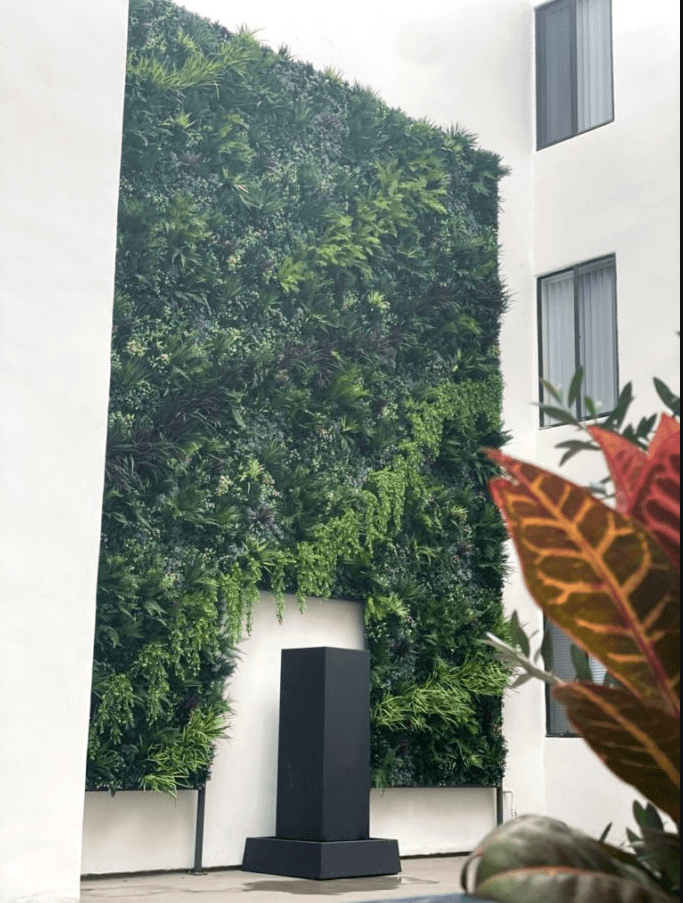 A lush Bespoke Pattern Artificial Green Wall in Los Angeles covers a wall between two white buildings, displaying a variety of green plants. In front of the wall, a tall black rectangular structure stands on a patio, with colorful leaves of a plant in the foreground.