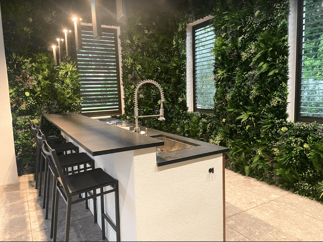 A modern kitchen in Florida features a long black island counter, four bar stools, and a sleek metal faucet. Residential artificial green wall panels in Orlando add lush greenery, while large windows with horizontal slats flood the space with natural light.