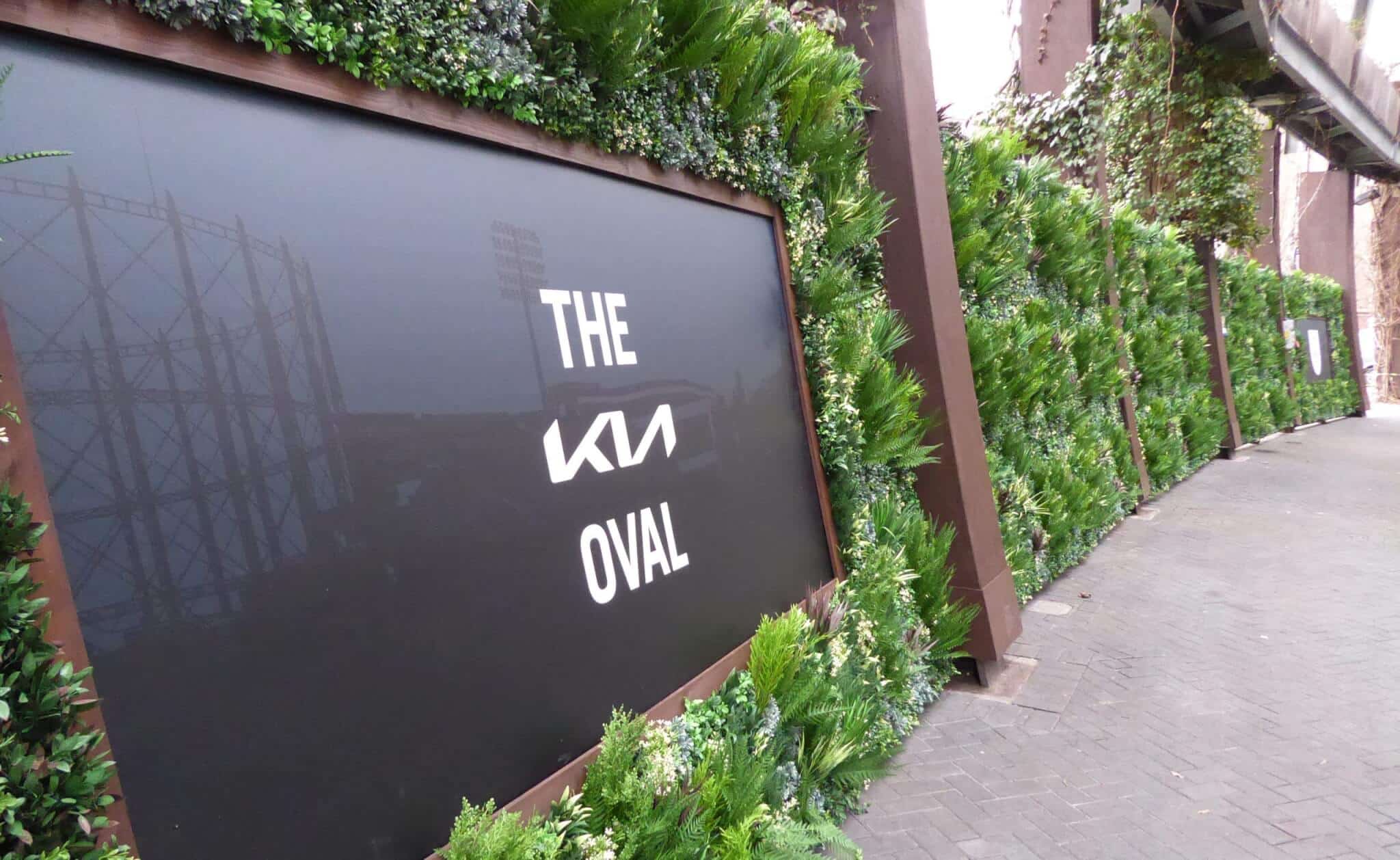 A large black sign with white text reads The Oval next to a logo. It is surrounded by lush green plants, forming a vertical garden. The pathway beside it is paved with stones.