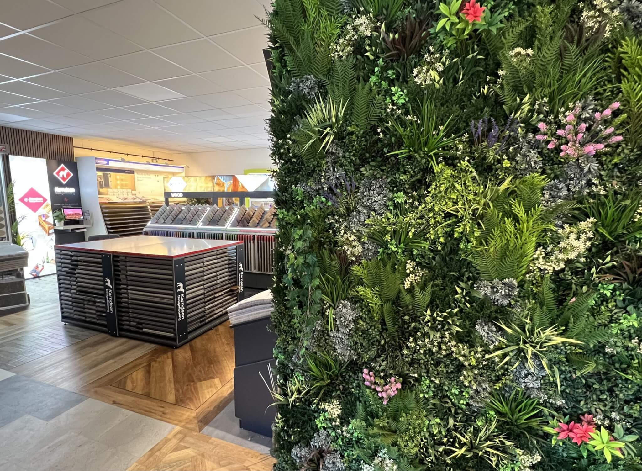 A showroom corner featuring a lush green vertical garden wall with various plants and flowers. In the background, there are displays with tiles and flooring samples under bright ceiling lights.