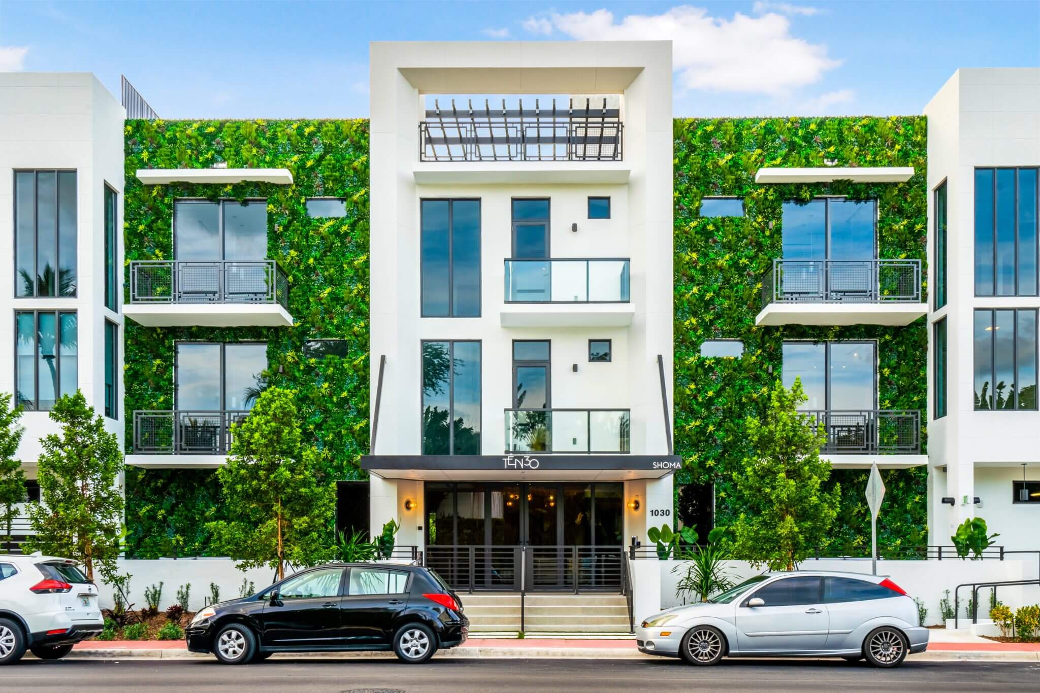 A modern apartment building in Miami boasts a lush exterior residential green wall on its facade. It features large windows and balconies, with cars parked out front on the Florida street under a clear sky dotted with a few clouds.