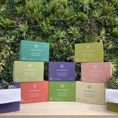 Exhibit of Large Grasses Texture Box packages in a variety of colors set out on a wooden surface, with a lush green vertical garden in the background that elevates the display.