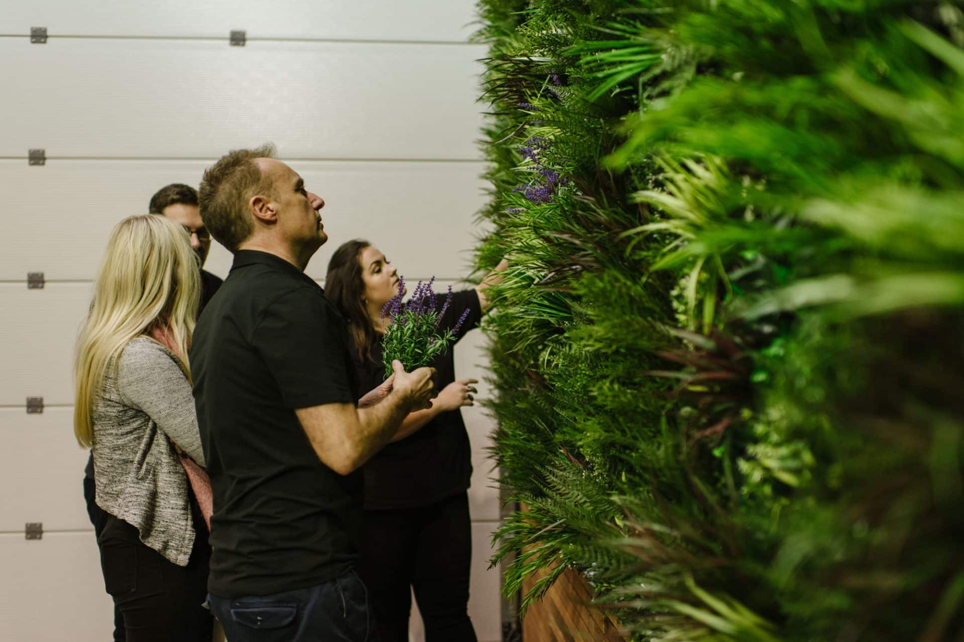 A group of four people, three standing together and one slightly apart, examines a lush vertical garden with diverse green foliage. The setting appears to be indoors, with a garage-style door visible in the background.