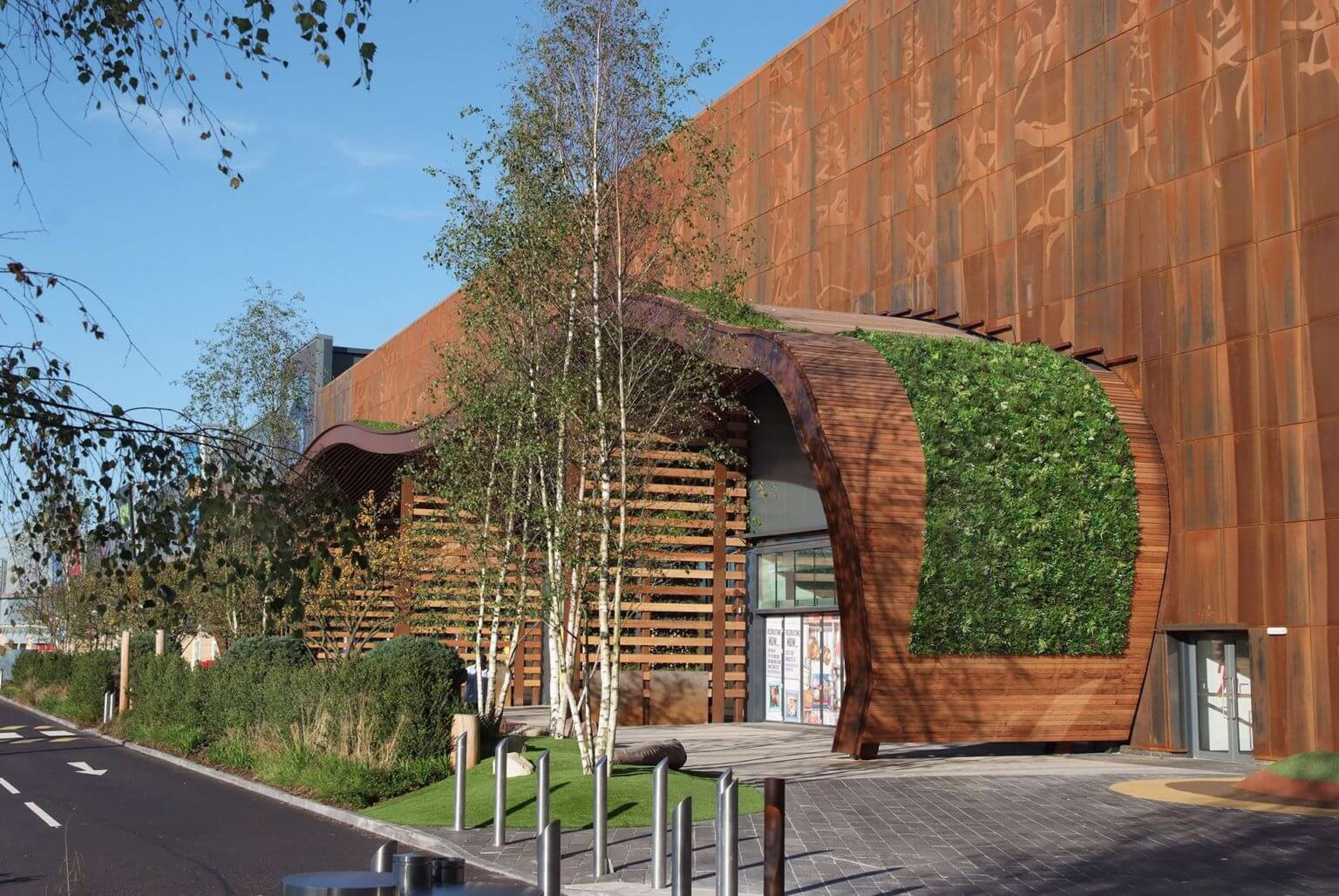 A modern building featuring a rust-colored metal facade and wooden accents boasts an artificial green wall that enhances its entrance. Trees and shrubs add greenery to the pathway beside a road, creating an eco-friendly atmosphere under a clear blue sky.