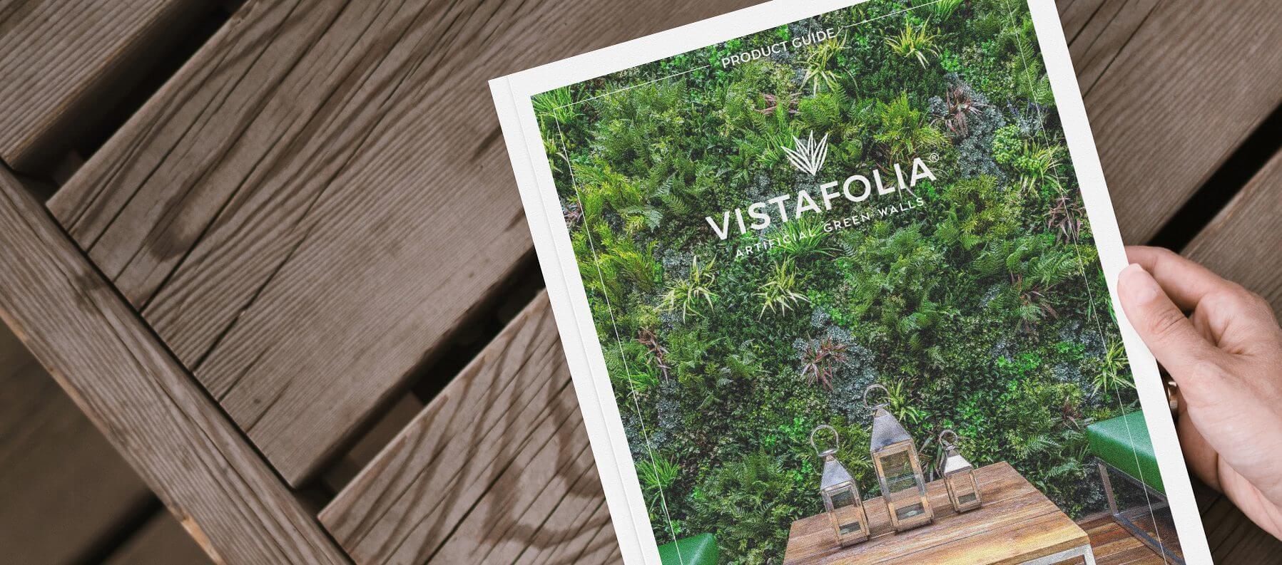 A person holds a Vistafolia Artificial Green Wall Panels product guide, featuring an image of a lush green vertical garden on the cover, with wooden steps visible in the background.