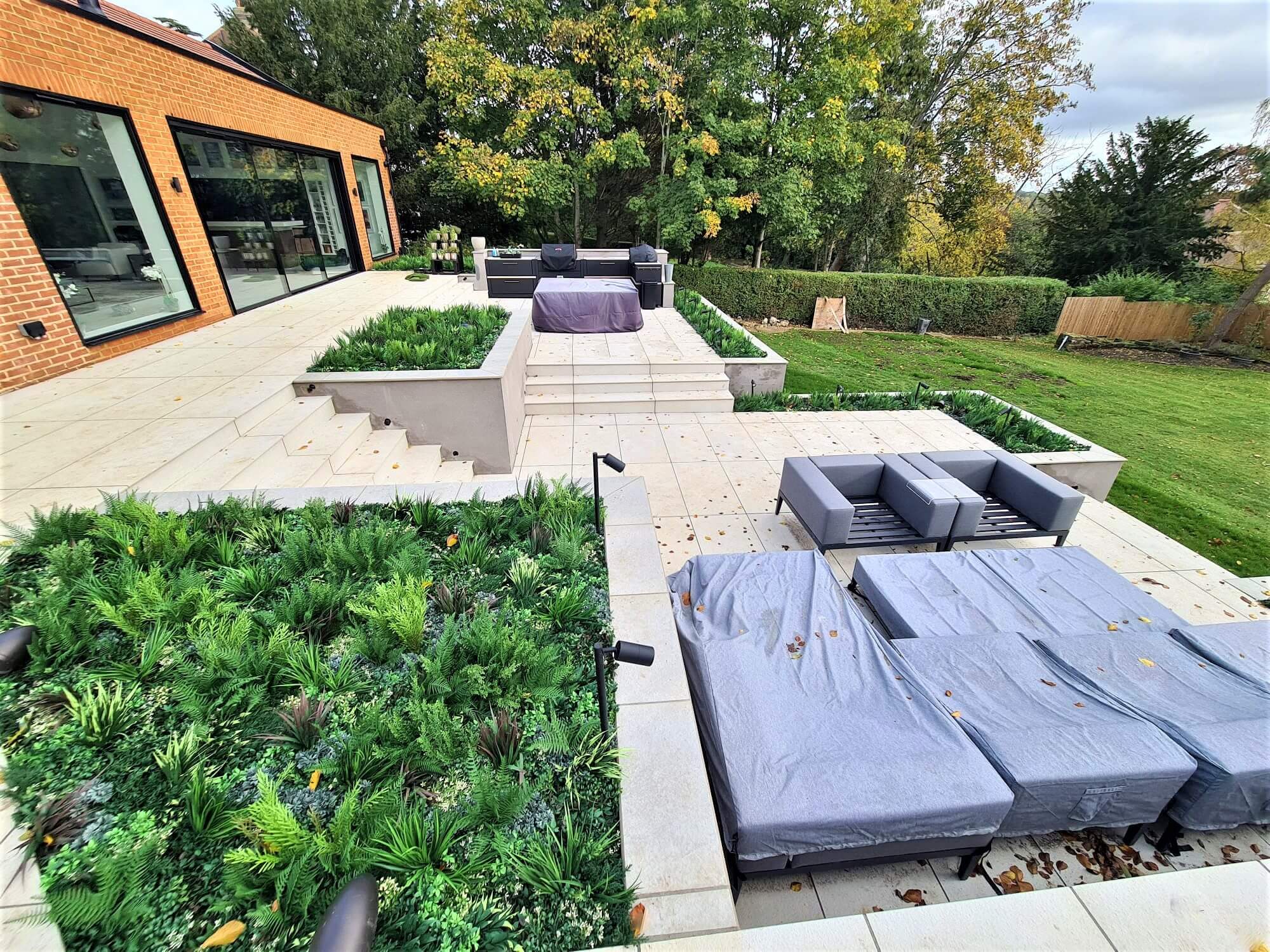 Modern patio with gray sofas and tables, surrounded by lush green plants. The multi-tiered design features beige stone tiles and steps, leading to a grassy lawn. Trees and a hedge line the background, under a cloudy sky.