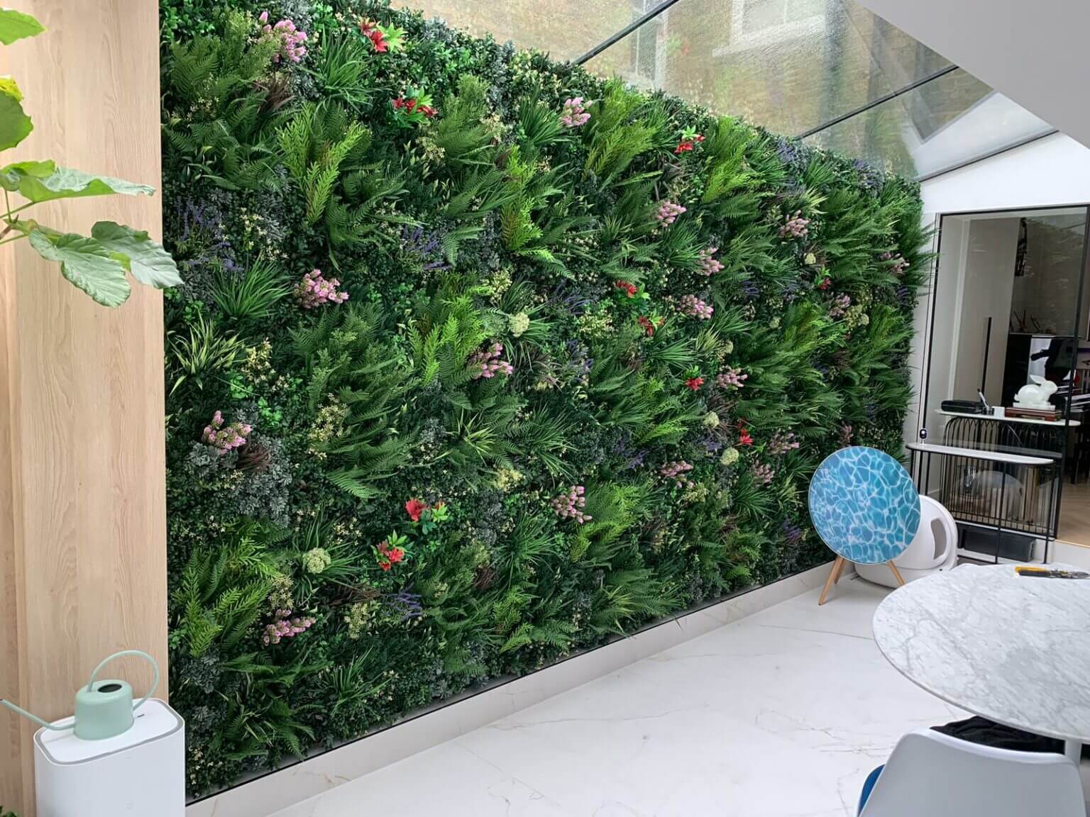 A modern interior showcases a lush vertical garden wall, blending artificial plants with vibrant pink and red flowers. The space includes a round blue chair, a white round table, and a wooden watering can on a tiled floor, emphasizing the importance of picking the right materials for realism.